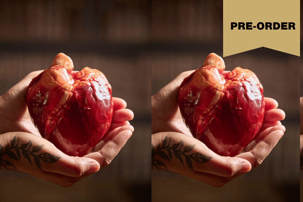 A split-screen lifestyle image showing a person's tattooed hands holding a realistic red and orange anatomical heart made of chocolate. The background is dark and blurred, and a gold 'PRE-ORDER' banner is in the top right corner.