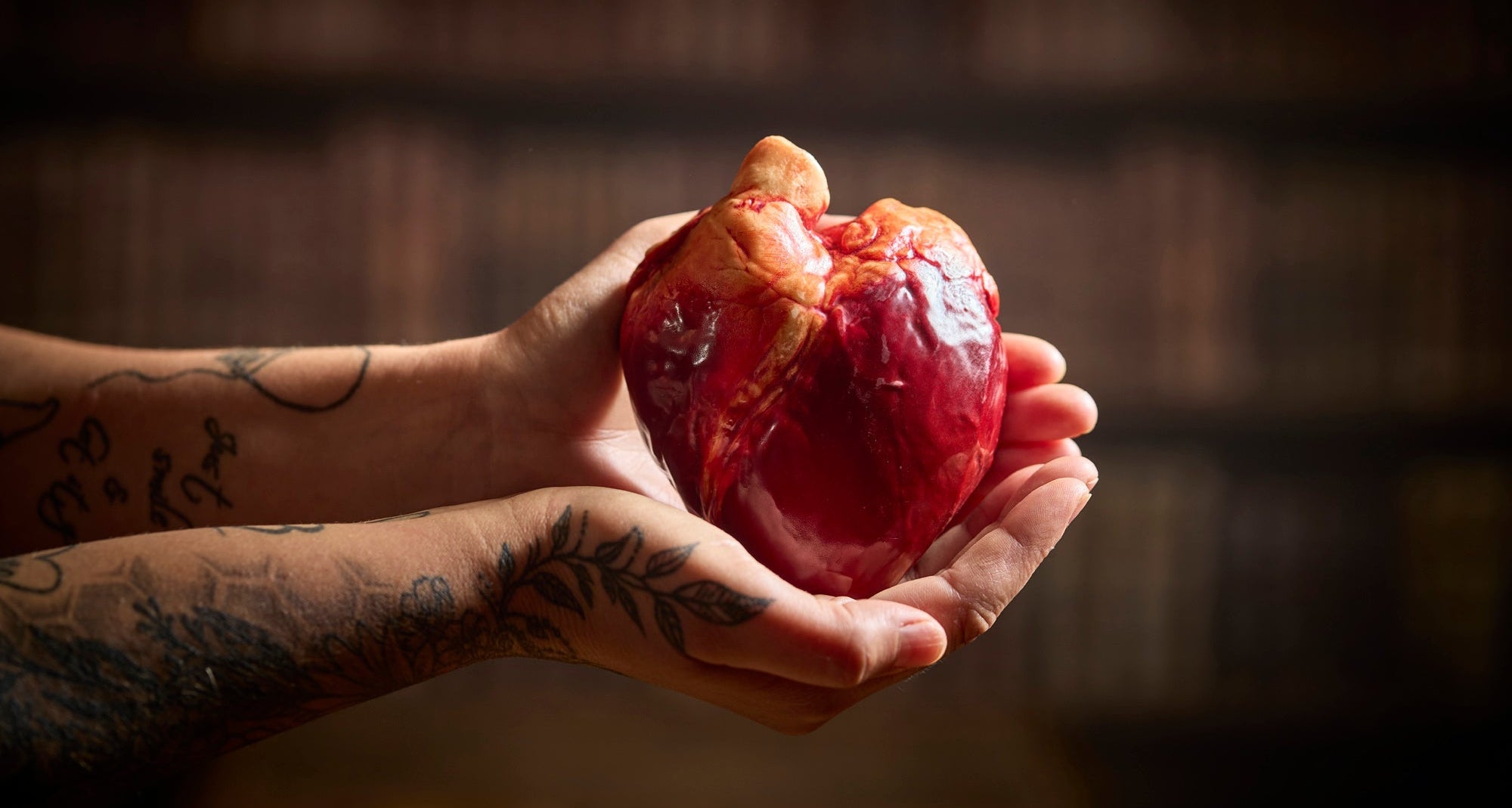 A pair of tattooed hands holding a realistic heart in red made of chocolate against a dark blurred background.