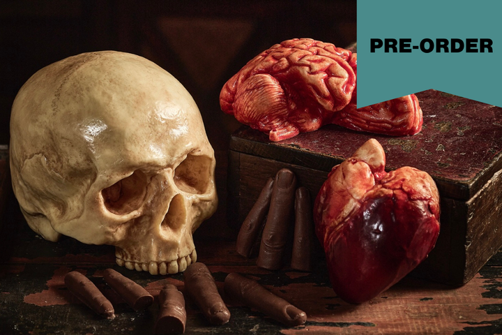 A still life arrangement featuring chocolate sculptures of realistic human skull, a brain, a heart, and several fingers on a dark wooden surface, with a teal corner label reading “PRE-ORDER.”.